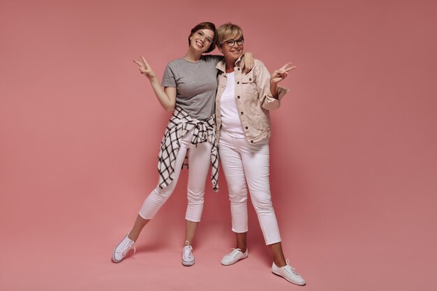 Две прекрасные женщины с короткими волосами и в современных очках в белых узких брюках и легких кроссовках улыбаются и демонстрируют знаки мира на розовом фоне.