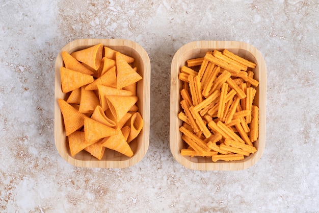 Two wooden bowls of triangle chips and breadsticks on a stone surface.