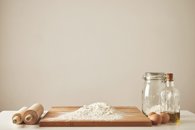 Two wooden rolling pins, extra virgin olive oil, transparent jar and wooden cutting board with white flour, chiken eggs isolated. Everything prepared for dough making