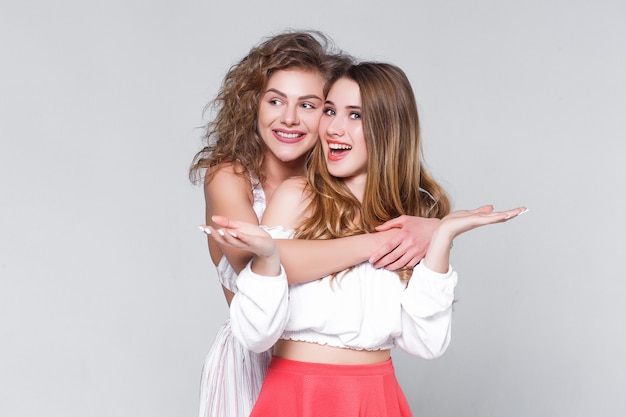 Two young beautiful blond smiling girls hugging in trendy summer clothes. Carefree women isolated on gray background.