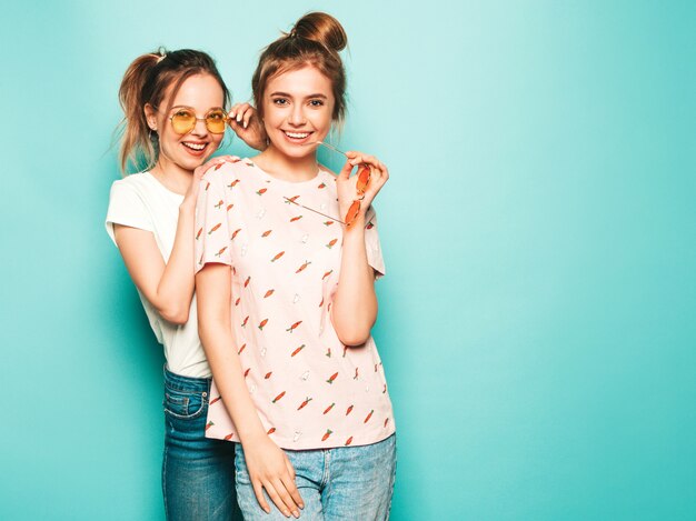 Две молодые красивые белокурые улыбающиеся хипстерские девочки в модных летних хипстерских джинсовых одеждах. Сексуальные беззаботные женщины позируют возле синей стены. Модные и позитивные модели с удовольствием в солнцезащитных очках