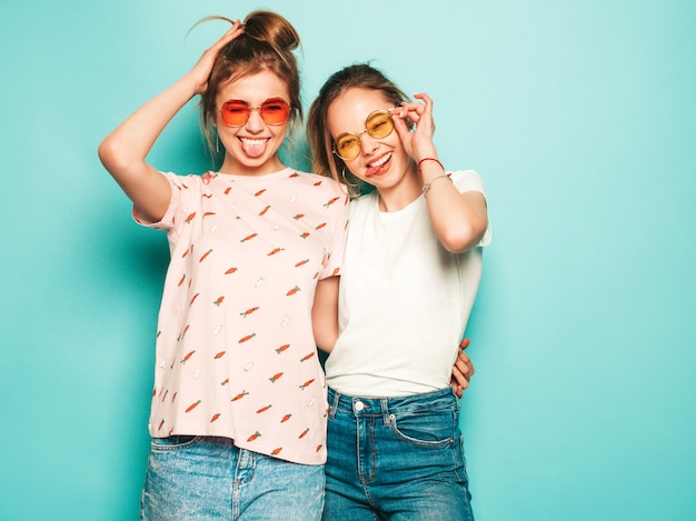 Two young beautiful blond smiling hipster girls in trendy summer hipster jeans clothes. sexy carefree women posing near blue wall. trendy and positive models having fun in sunglasses