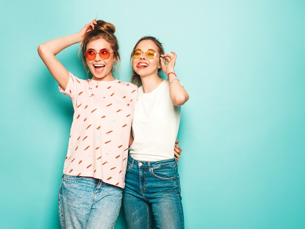 Two young beautiful blond smiling hipster girls in trendy summer hipster jeans clothes. sexy carefree women posing near blue wall. trendy and positive models having fun in sunglasses