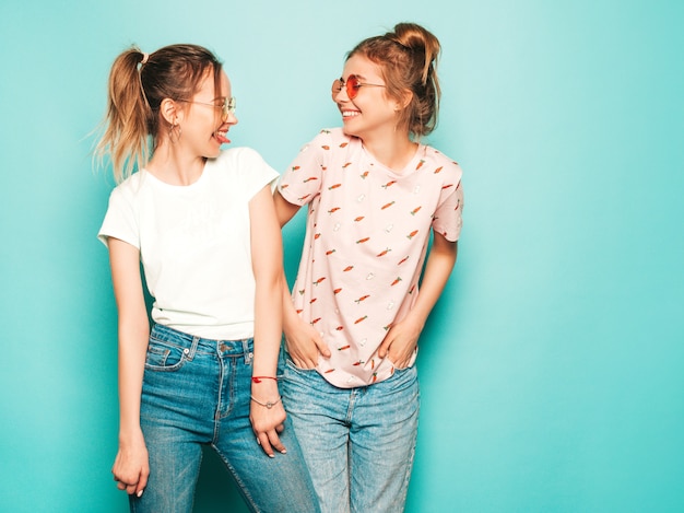 Two young beautiful blond smiling hipster girls in trendy summer hipster jeans clothes. Sexy carefree women posing near blue wall. Trendy and positive models having fun in sunglasses