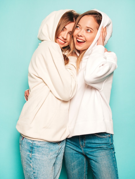 Two young beautiful blond smiling hipster girls in trendy summer hoodie clothes. sexy carefree women posing near blue wall. trendy and positive models having fun