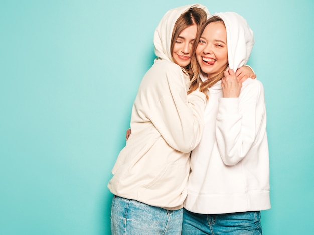 Two young beautiful blond smiling hipster girls in trendy summer hoodie clothes. sexy carefree women posing near blue wall. trendy and positive models show tongue sign in sunglasses