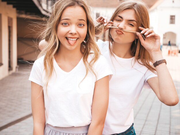Две молодые красивые белокурые улыбающиеся хипстерские девочки в модной летней белой футболке одеваются. Позитивные модели с удовольствием. Делаем усы с волосами и показываем язык
