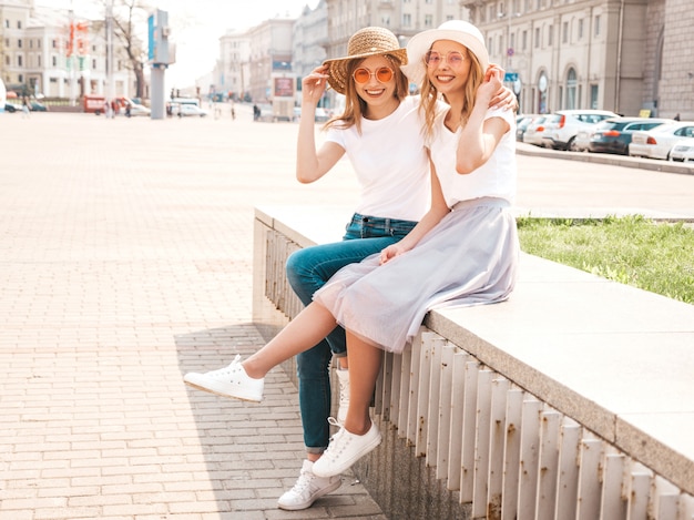 Due giovani belle ragazze sorridenti bionde dei pantaloni a vita bassa in vestiti bianchi alla moda della maglietta di estate. donne spensierate sexy che si siedono sul fondo della via.