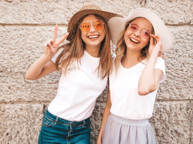 Two young beautiful blond smiling hipster girls in trendy summer white t-shirt clothes. Women posing in the street near wall. 