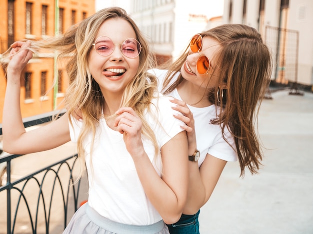 Two young beautiful blond smiling hipster girls in trendy summer white t-shirt clothes. women posing on street . Positive models having fun in sunglasses.Shows peace sign