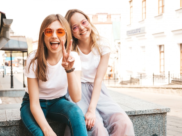 トレンディな夏の2人の若い美しいブロンド笑顔流行に敏感な女の子白いtシャツ服。通りに座っている女性。サングラスで楽しんでいる肯定的なモデル。ピースサインを示しています