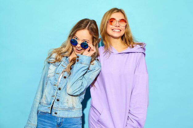 Two young beautiful blond smiling hipster women in trendy summer clothes. Sexy carefree women posing near blue wall in sunglasses. Positive models going crazy and hugging