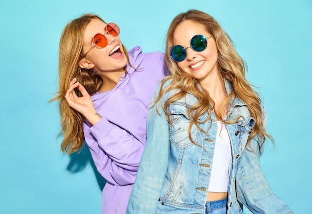 Две молодые красивые белокурые улыбающиеся женщины битник в модной летней одежды. Сексуальные беззаботные женщины позируют возле синей стене в солнцезащитные очки. Позитивные модели сходят с ума и обнимаются