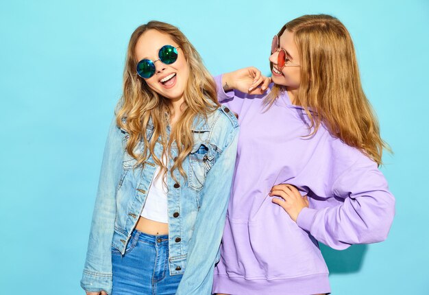 Two young beautiful blond smiling hipster women in trendy summer clothes. Sexy carefree women posing near blue wall in sunglasses. Positive models going crazy