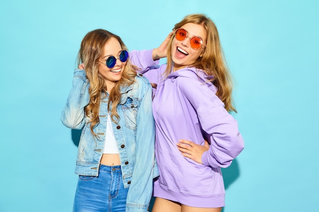 Two young beautiful blond smiling hipster women in trendy summer clothes. Sexy carefree women posing near blue wall in sunglasses. Positive models going crazy