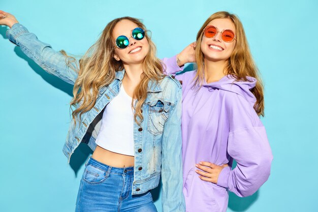 Two young beautiful blond smiling hipster women in trendy summer clothes. Sexy carefree women posing near blue wall in sunglasses. Positive models going crazy