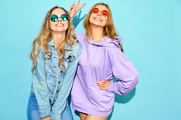 Две молодые красивые белокурые улыбающиеся женщины битник в модной летней одежды. Сексуальные беззаботные женщины позируют возле синей стене в солнцезащитные очки. Позитивные модели сходят с ума