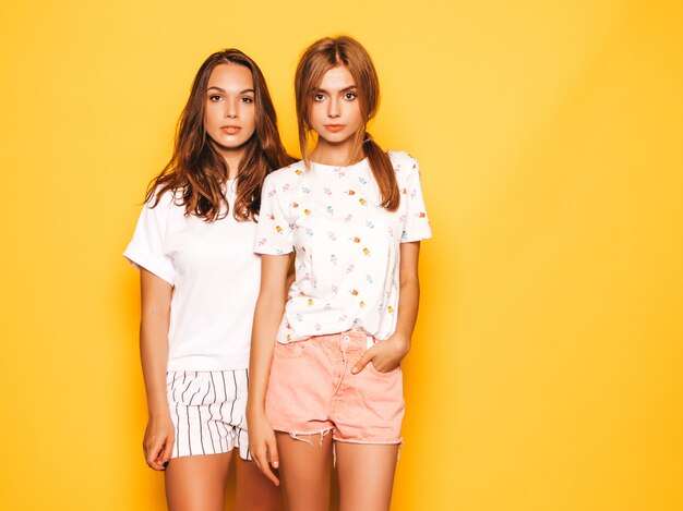 Two young beautiful boring hipster girls in trendy summer clothes. Sexy carefree women posing near yellow wall