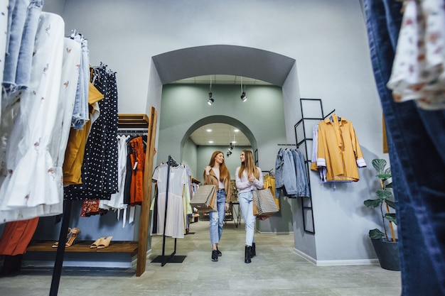 Free photo two young, beautiful girlfriends are walking in a style of loft showroom stylish things with gift bags, smiling at each other.