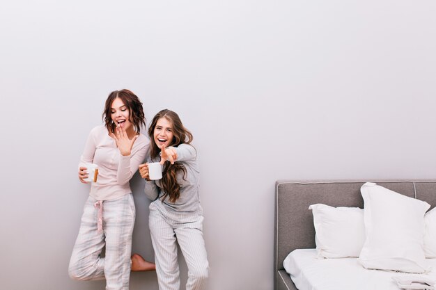 Two young beautiful girls in night pajamas with cups having fun  in sleeping room on grey wall . They look enjoyed and smiling.