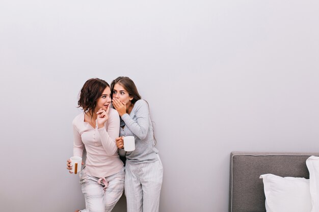 Two young beautiful girls in pajamas with cups having fun in sleeping room on grey wall . They whispering secrets to each other.