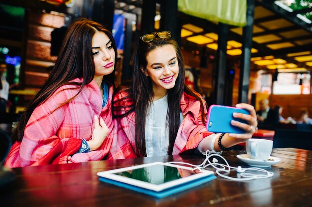 Two young and beautiful girls sitting at the table and doing selfie in the cafe