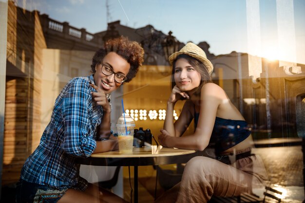 Две молодые красивые девушки улыбаются, говоря, отдыхая в кафе. Снято снаружи.