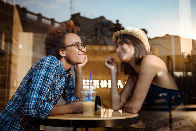 Two young beautiful girls smiling, speaking, resting in cafe. Shot from outside.