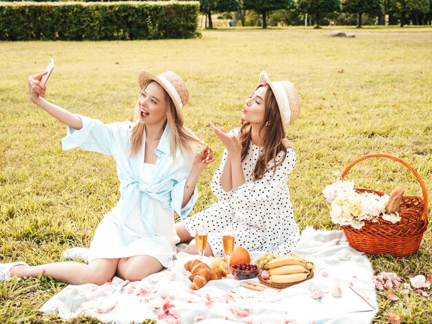 Two young beautiful hipster woman in trendy summer sundress and hats. Carefree women making picnic outside. 