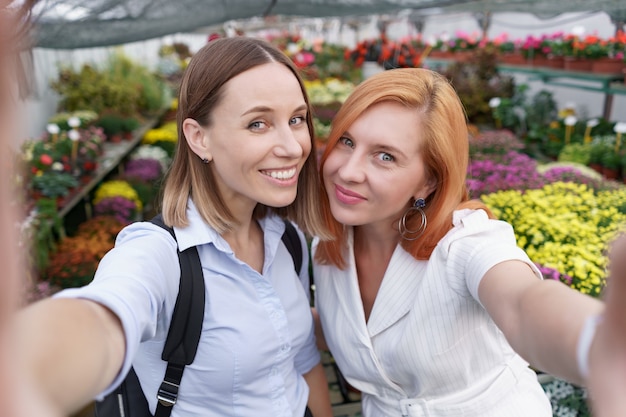 Two young beautiful ladies making selfie on flowers background in the greenhouse