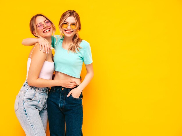 トレンディな夏の服を着た2人の若い美しい笑顔のブロンドのヒップスターの女性スタジオで黄色の壁の近くでポーズをとるセクシーな屈託のない女性
