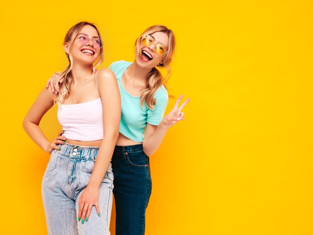 트렌디한 여름 옷을 입은 두 젊은 아름다운 웃는 금발 힙스터 여성 스튜디오의 노란색 벽 근처에서 포즈를 취하는 섹시하고 평온한 여성 긍정적인 모델 재미 쾌활하고 행복 선글라스
