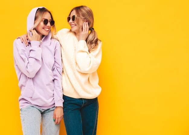 Two young beautiful smiling blond hipster female in trendy summer clothes Sexy carefree women posing near yellow wall in studio Positive models having fun Cheerful and happy In sunglasses