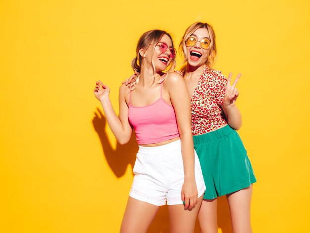 Two young beautiful smiling blond hipster female in trendy summer clothes Sexy carefree women posing near yellow wall in studio Positive models having fun Cheerful and happy In sunglasses