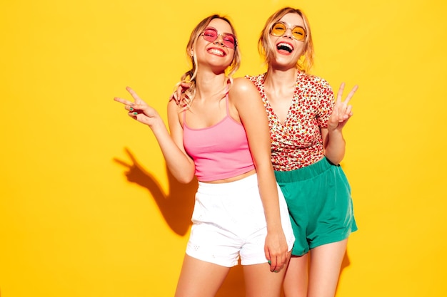 Two young beautiful smiling blond hipster female in trendy summer clothes Sexy carefree women posing near yellow wall in studio Positive models having fun Cheerful and happy In sunglasses