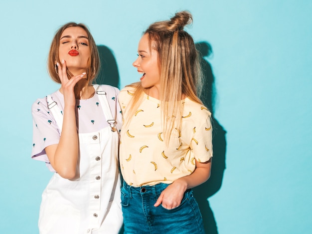 Две молодые красивые улыбающиеся белокурые хипстерские девочки в модной летней красочной футболке одеваются. и дает поцелуй