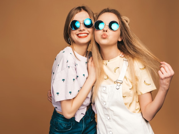 トレンディな夏のカラフルなtシャツの服の2人の若い美しい笑顔金髪流行に敏感な女の子。丸いサングラスでベージュ色の背景にポーズセクシーな屈託のない女性。楽しいポジティブモデル