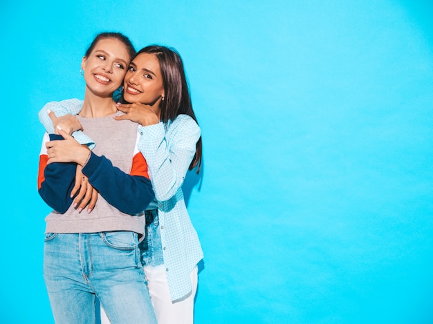 Две молодые красивые улыбающиеся белокурые хипстерские девочки в модной летней красочной футболке одеваются. Сексуальные беззаботные женщины позируют возле синей стены. Веселые позитивные модели