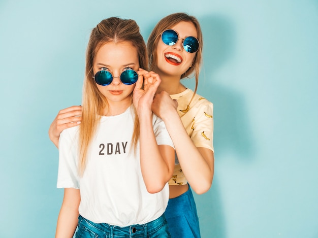 Due giovani belle ragazze bionde sorridenti dei pantaloni a vita bassa in vestiti variopinti della maglietta di estate alla moda.