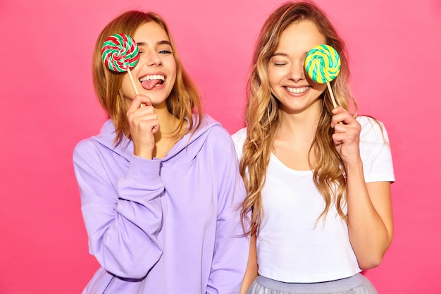 Two young beautiful smiling blond hipster women in trendy summer clothes. Carefree hot women posing near pink wall. Positive models cover eyes by lollipop