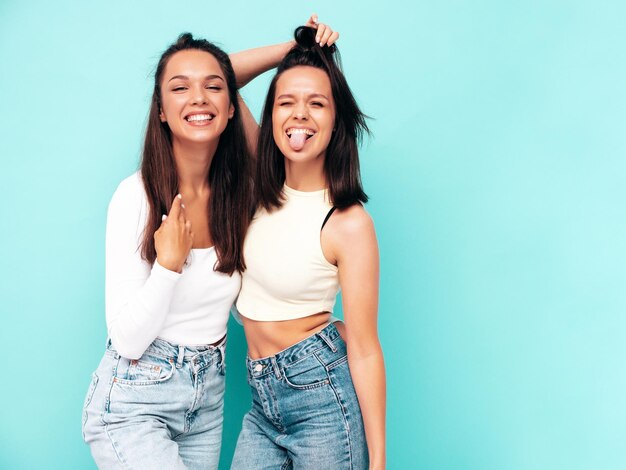 Two young beautiful smiling brunette hipster female in trendy summer clothes Sexy carefree women posing near blue wall Positive models having fun Cheerful and happy