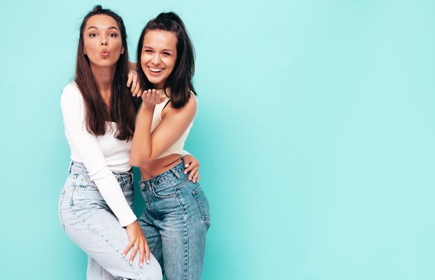 Two young beautiful smiling brunette hipster female in trendy summer clothes Sexy carefree women posing near blue wall Positive models having fun Cheerful and happy