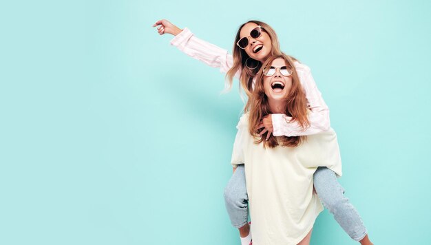 Two young beautiful smiling brunette hipster female in trendy summer clothes Sexy carefree women posing near blue wall Positive models having fun in sunglasses Gives piggyback riding