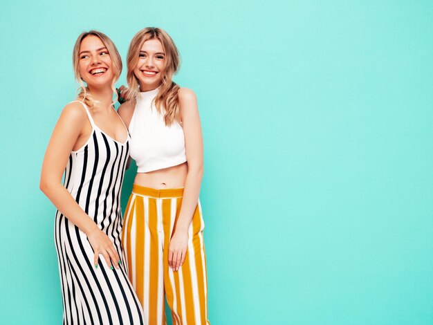 트렌디한 여름 옷을 입은 두 젊은 아름다운 웃는 갈색 머리 힙스터 여성 스튜디오의 파란색 벽 근처에서 포즈를 취하는 섹시한 평온한 여성 긍정적인 모델 재미 명랑하고 행복