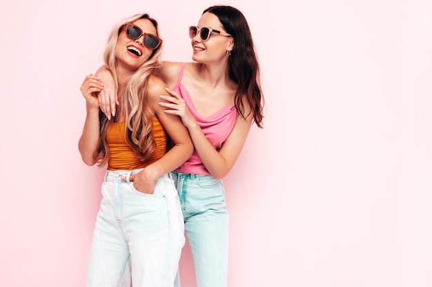 Two young beautiful smiling brunette hipster female in trendy summer clothes Sexy carefree women posing near pink wall Positive models having fun Cheerful and happy in sunglasses
