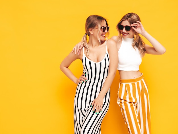 Две молодые красивые улыбающиеся брюнетки-хипстеры в модной летней одежде Сексуальные беззаботные женщины позируют возле желтой стены в студии Позитивные модели веселятся Веселые и счастливые В солнцезащитных очках