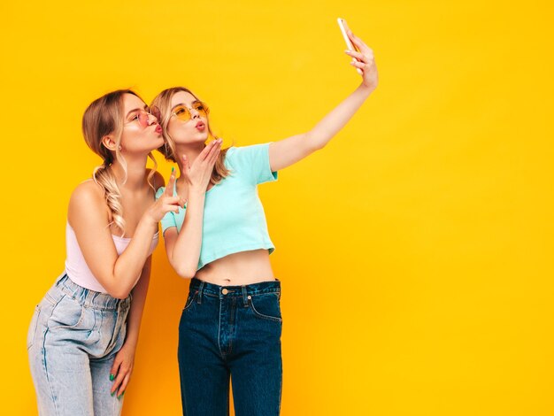 Two young beautiful smiling brunette hipster female in trendy summer clothes Sexy carefree women posing near yellow wall in studio Positive models having fun Cheerful and happy Taking selfie