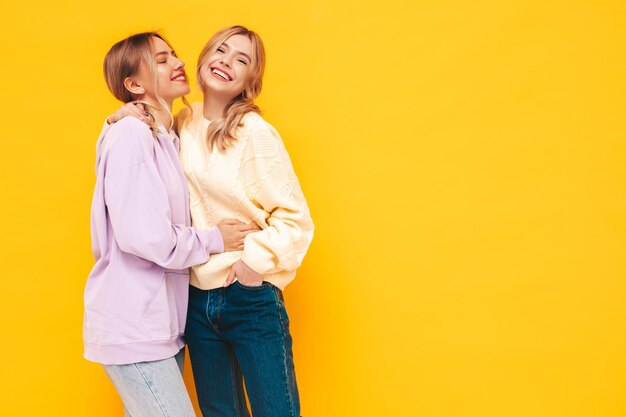 트렌디한 여름 옷을 입은 두 젊은 아름다운 웃는 갈색 머리 힙스터 여성 스튜디오의 노란색 벽 근처에서 포즈를 취하는 섹시하고 평온한 여성 긍정적인 모델 재미 명랑하고 행복