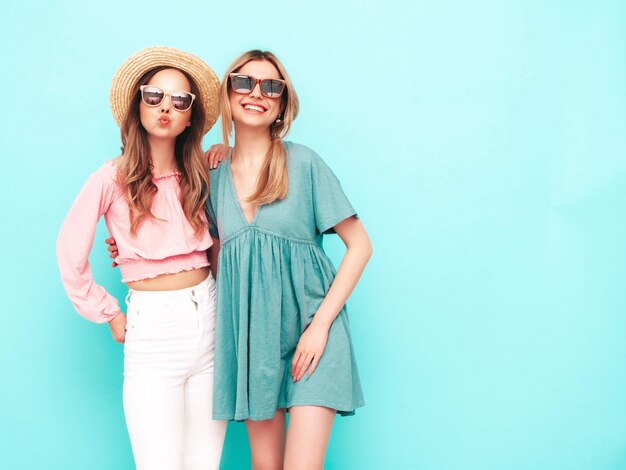 최신 유행의 여름 드레스를 입은 두 젊은 아름다운 웃는 갈색 머리 힙스터 여성 파란색 벽 근처에서 포즈를 취하는 섹시하고 평온한 여성 긍정적인 모델은 재미 있고 쾌활하고 행복합니다. 모자와 선글라스