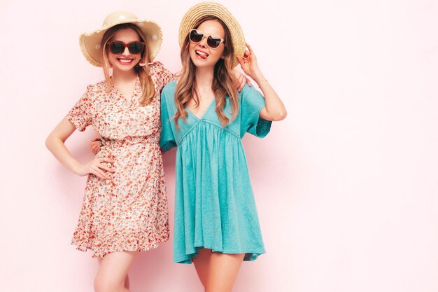 트렌디한 여름 드레스를 입은 두 젊은 아름다운 웃는 갈색 머리 힙스터 여성 분홍색 벽 근처에서 포즈를 취하는 섹시하고 평온한 여성 긍정적인 모델은 재미 있고 쾌활하고 행복합니다. 모자와 선글라스